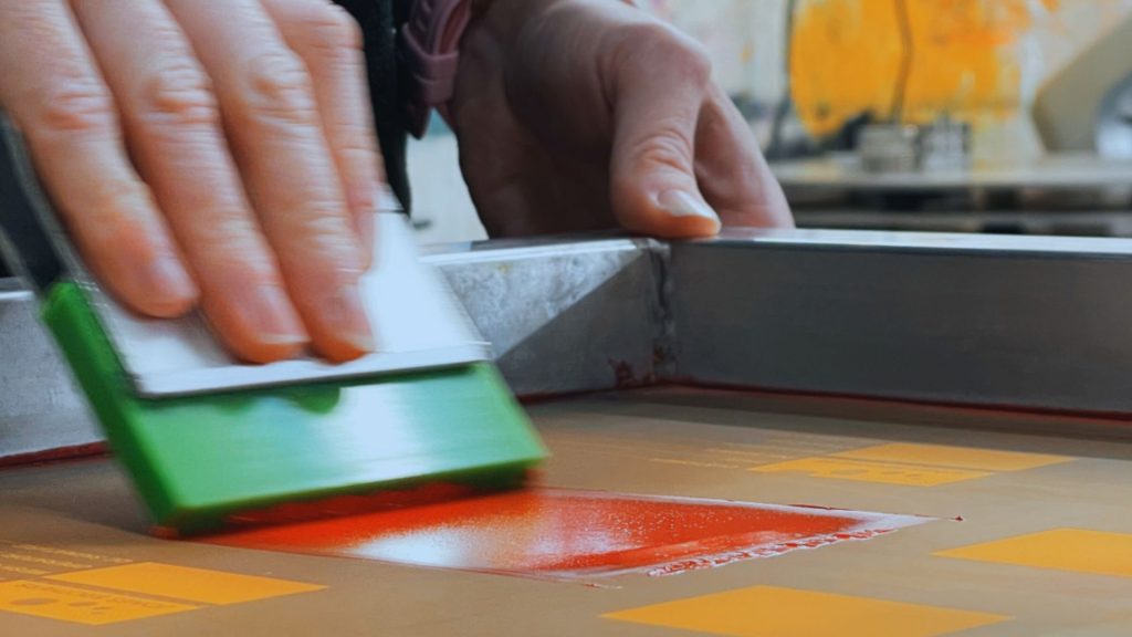 printer using squeegee to print colour through colour swatch screen to test the colour against the substrate that will be compared under a colour light box to ensure it is the same.