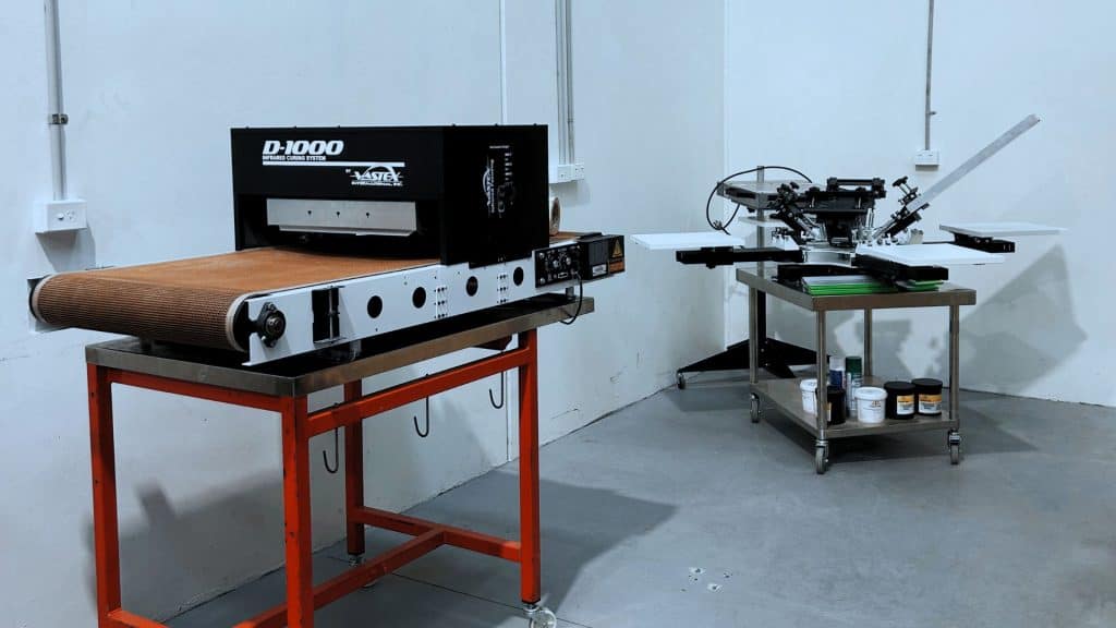 Chris Benson, self-taught screen printer and owner of Cherry Studios, pictured in his production space with screens and equipment behind him—capturing the hustle, growth and authenticity described in the Behind the Mesh December 2025 interview.