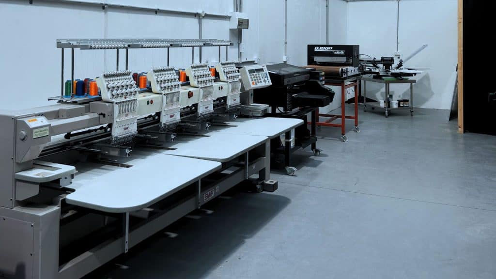 Chris Benson, self-taught screen printer and owner of Cherry Studios, pictured in his production space with screens and equipment behind him—capturing the hustle, growth and authenticity described in the Behind the Mesh December 2025 interview.