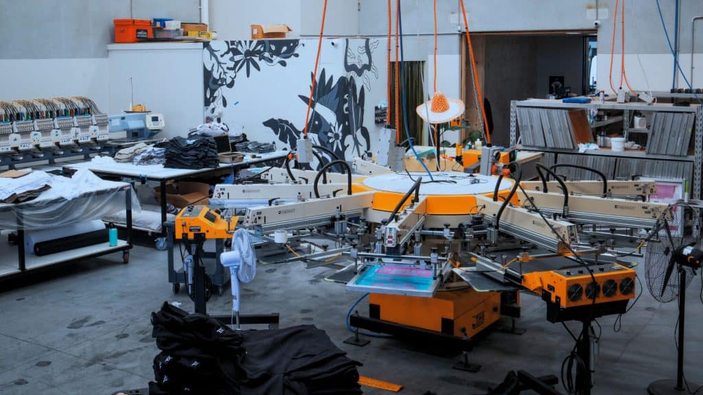 Chris Benson, self-taught screen printer and owner of Cherry Studios, pictured in his production space with screens and equipment behind him—capturing the hustle, growth and authenticity described in the Behind the Mesh December 2025 interview.