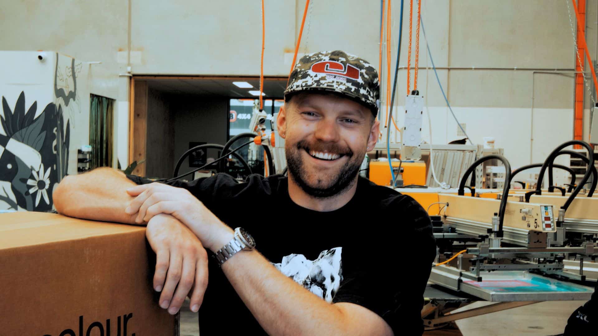 Chris Benson, self-taught screen printer and owner of Cherry Studios, pictured in his production space with screens and equipment behind him—capturing the hustle, growth and authenticity described in the Behind the Mesh December 2025 interview.
