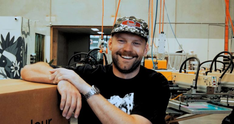Chris Benson, self-taught screen printer and owner of Cherry Studios, pictured in his production space with screens and equipment behind him—capturing the hustle, growth and authenticity described in the Behind the Mesh December 2025 interview.